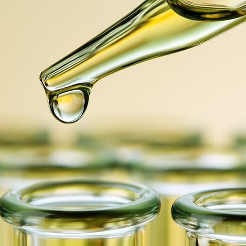 A close-up of a glass dropper holding a golden yellow liquid, with a single clear droplet hanging above the open tops of glass test tubes in the background.